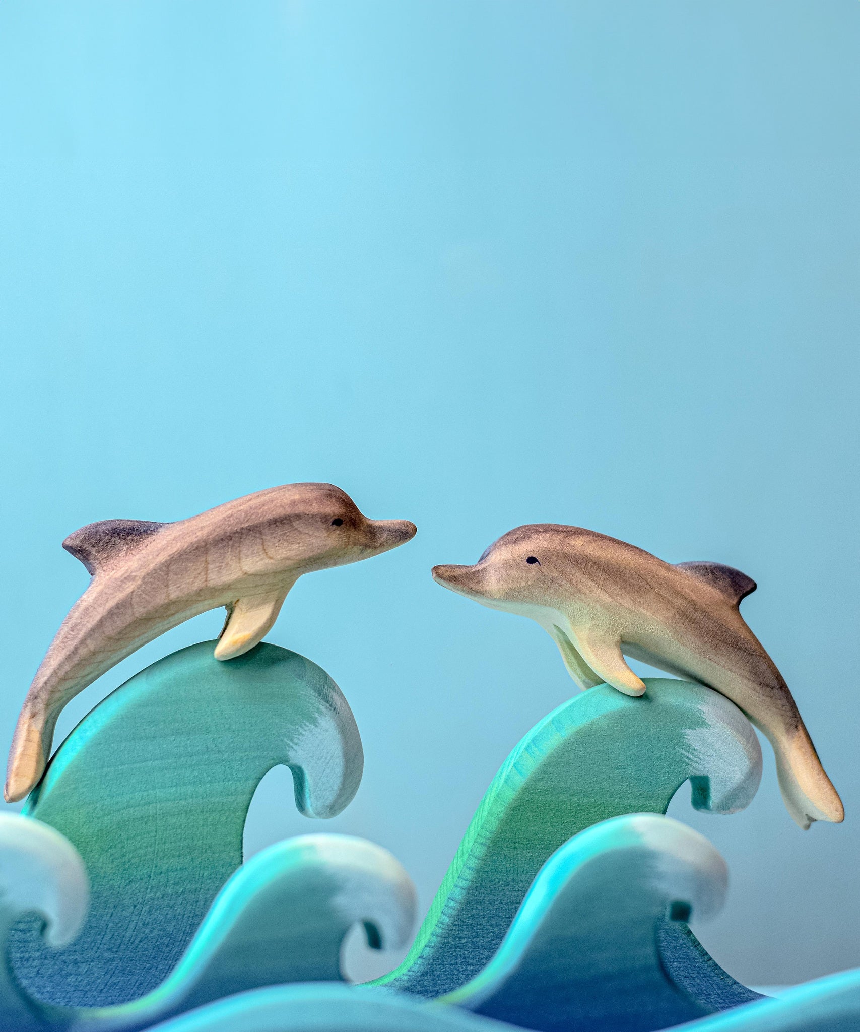 Two Bumbu Wooden Dolphins perched on waves in front of a blue background. 