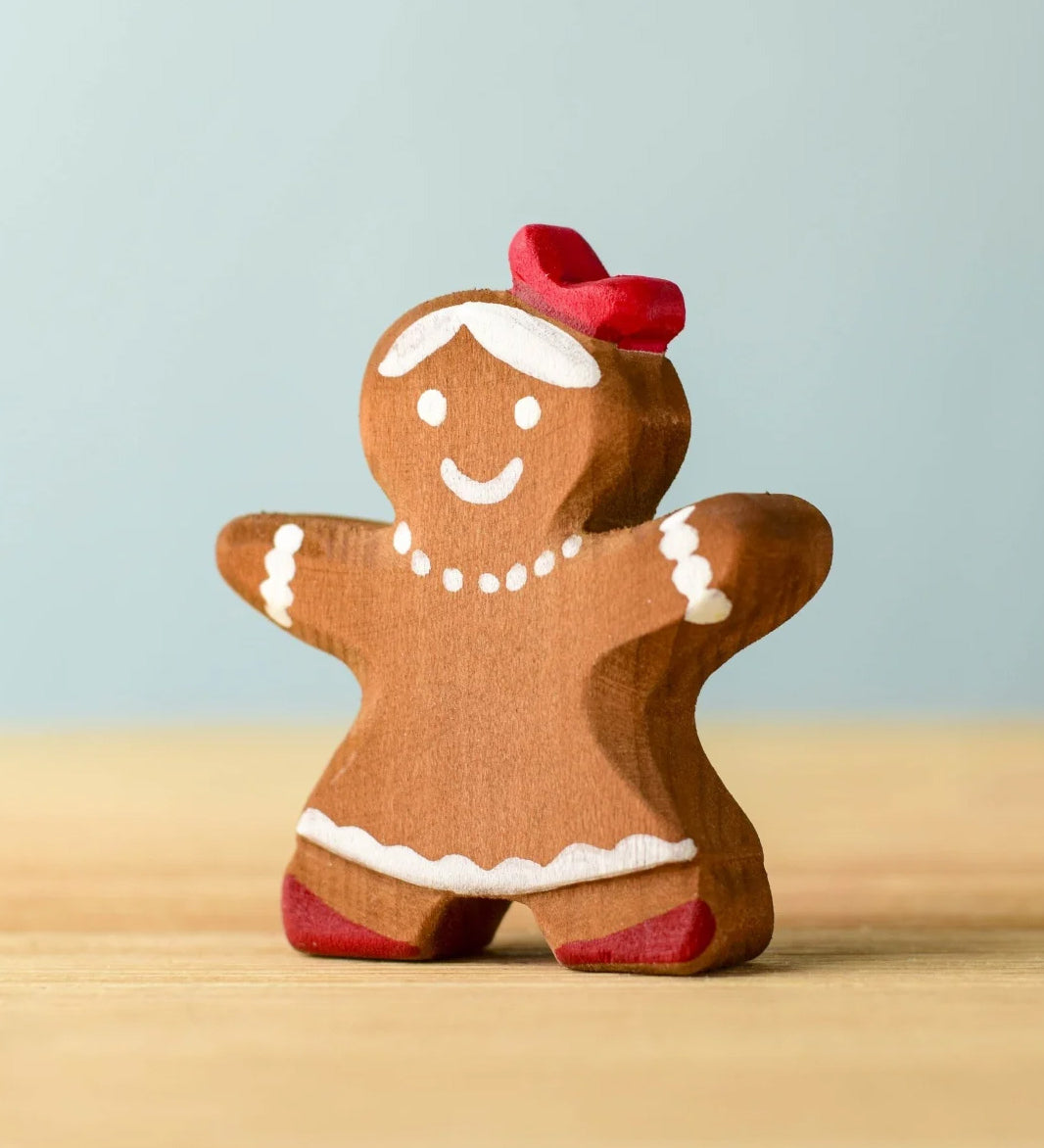 A wooden Bumbu gingerbread cookie girl with hand painted white detailing wearing a red bow on her head 