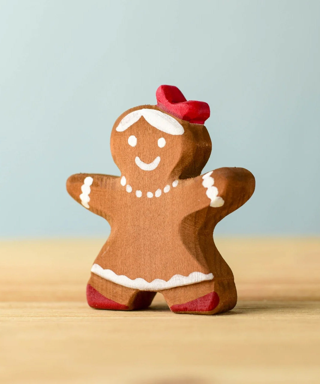 A wooden Bumbu gingerbread cookie girl with hand painted white detailing wearing a red bow on her head 