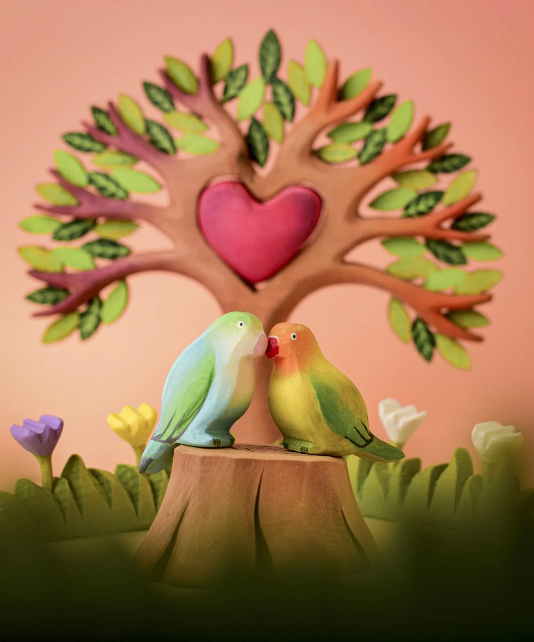 Bumbu wooden blue and green lovebird figure perched on a Bumbu wooden stump with the Bumbu heart in a tree set in the background.