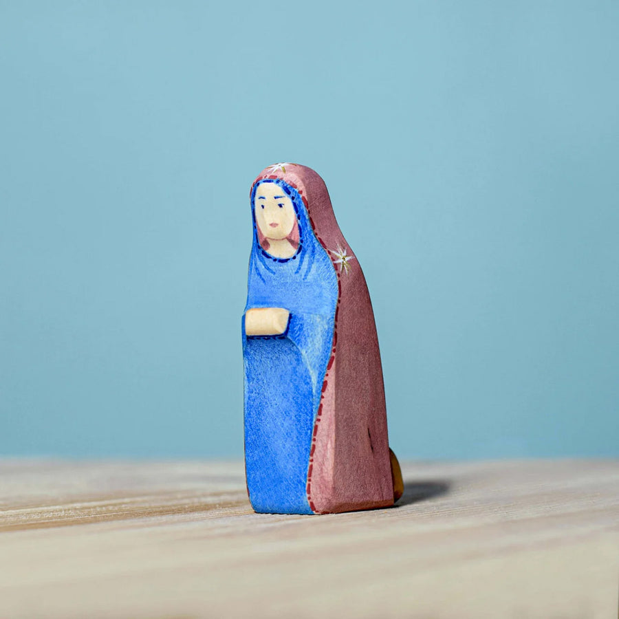 Bumbu Handmade Wooden Mary Figure kneeling down on a wooden table and blue background.