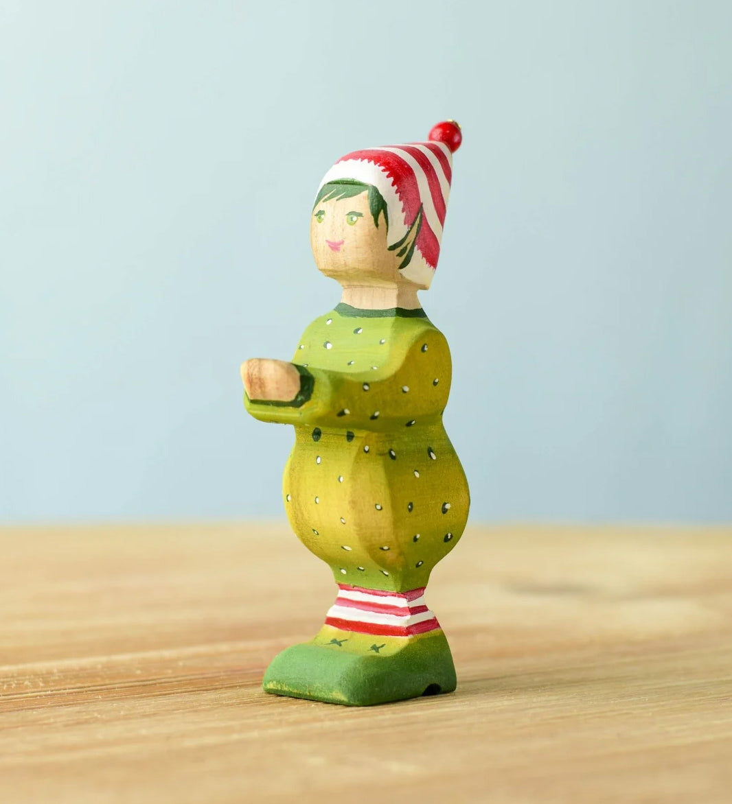 A wooden Bumbu Santas elf wearing a red and white striped hat and a green outfit with its hands holding out to carry presents.