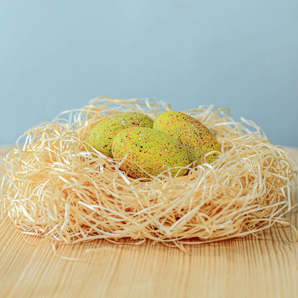 Bumbu Wooden Stegosaurus Eggs Set. Three speckled yellow dinosaur eggs sat in a nest. They are displayed on a wooden table and a blue background