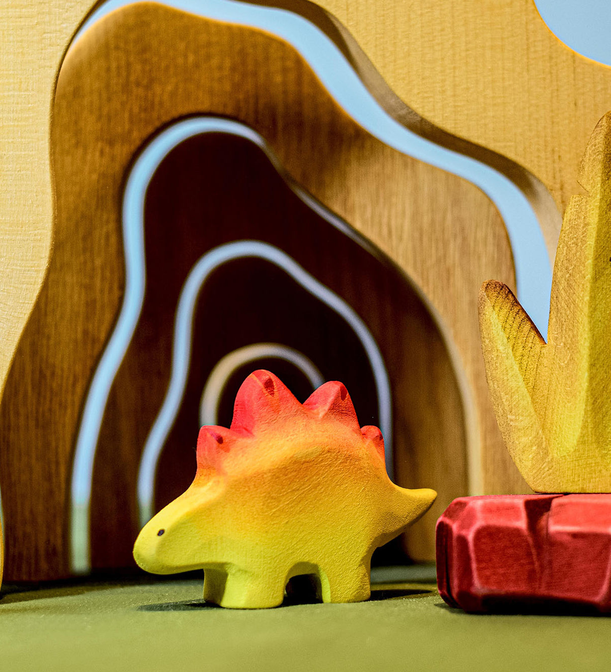 A wooden Bumbu baby stegosaurus stood in front of wooden cave arches and a red coloured wooden rock.