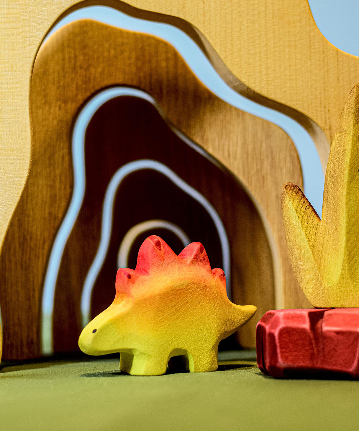 A wooden Bumbu baby stegosaurus stood in front of wooden cave arches and a red coloured wooden rock.