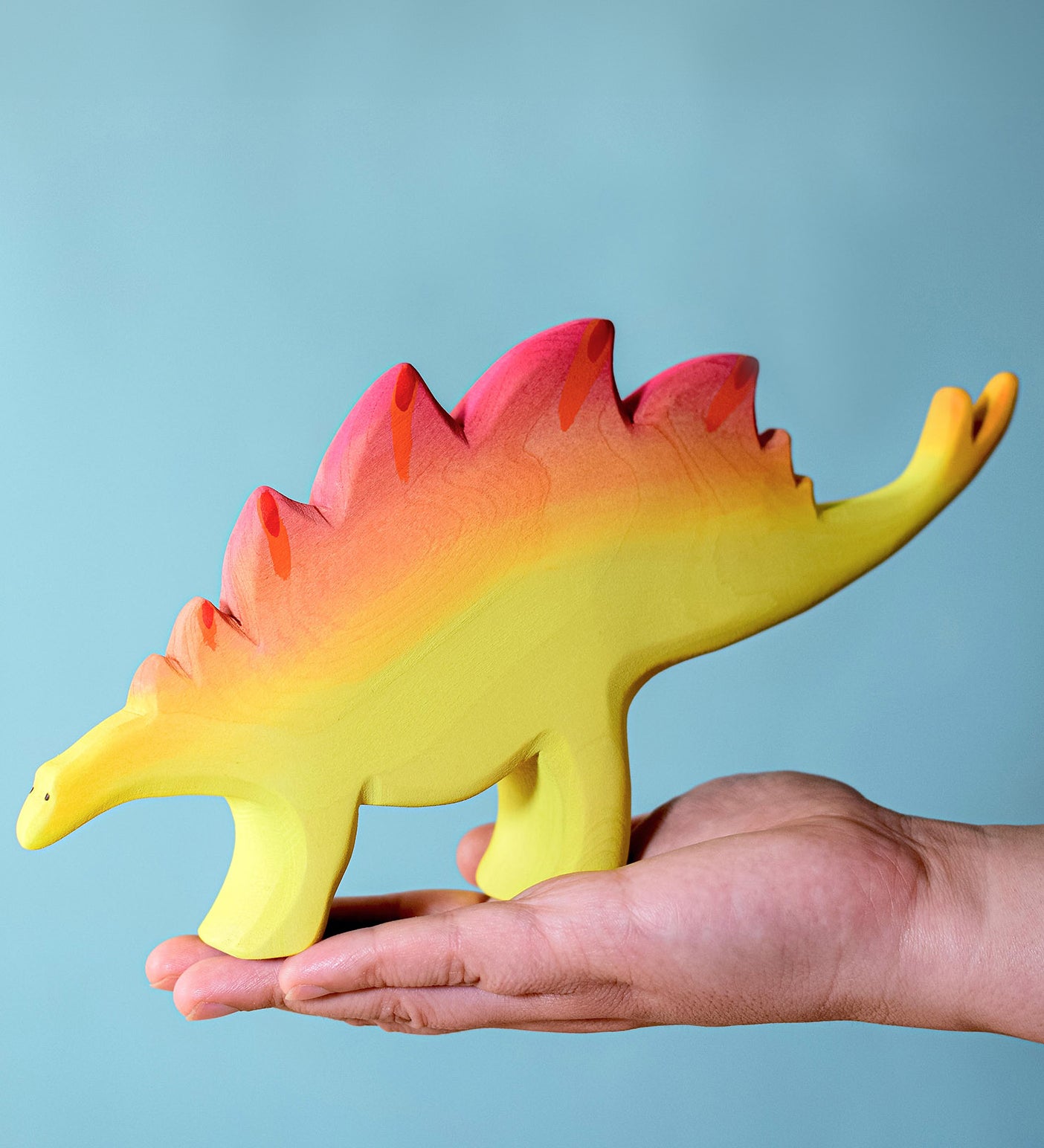 A large wooden Bumbu stegosaurus figure balancing on an adult's hand against a blue background.