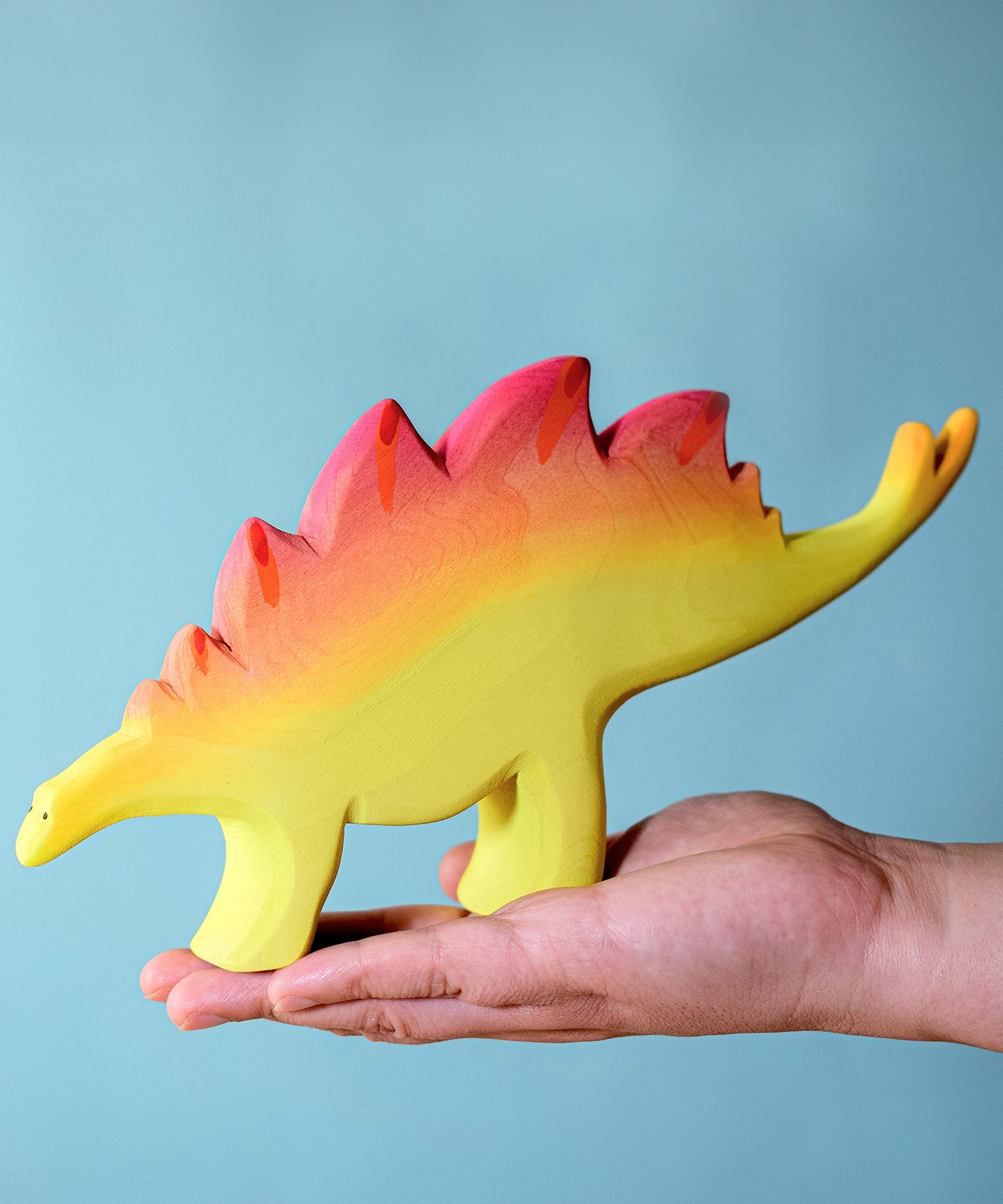 A large wooden Bumbu stegosaurus figure balancing on an adult's hand against a blue background.