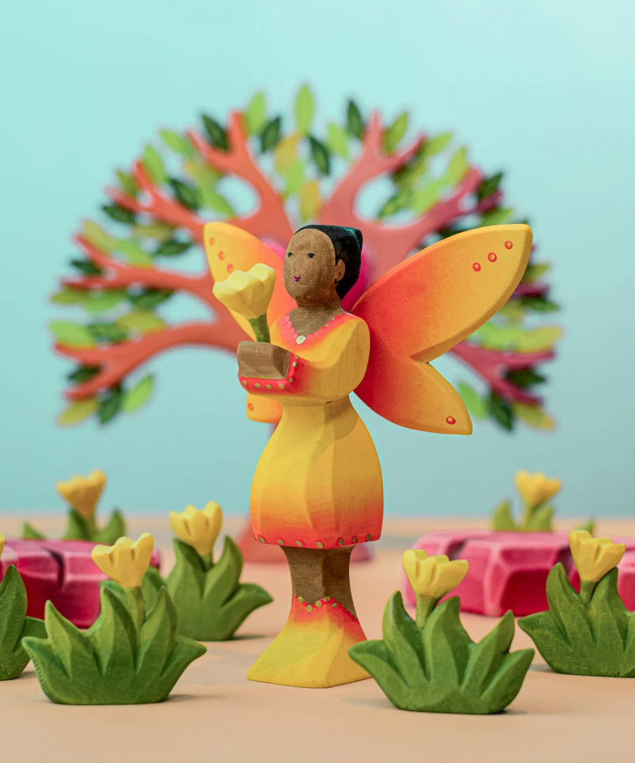 A Bumbu Wooden Sunflower Fairy holding a small yellow flower placed in a small world play scene surronded by yellow flowers with a wooden heart tree in the background. 