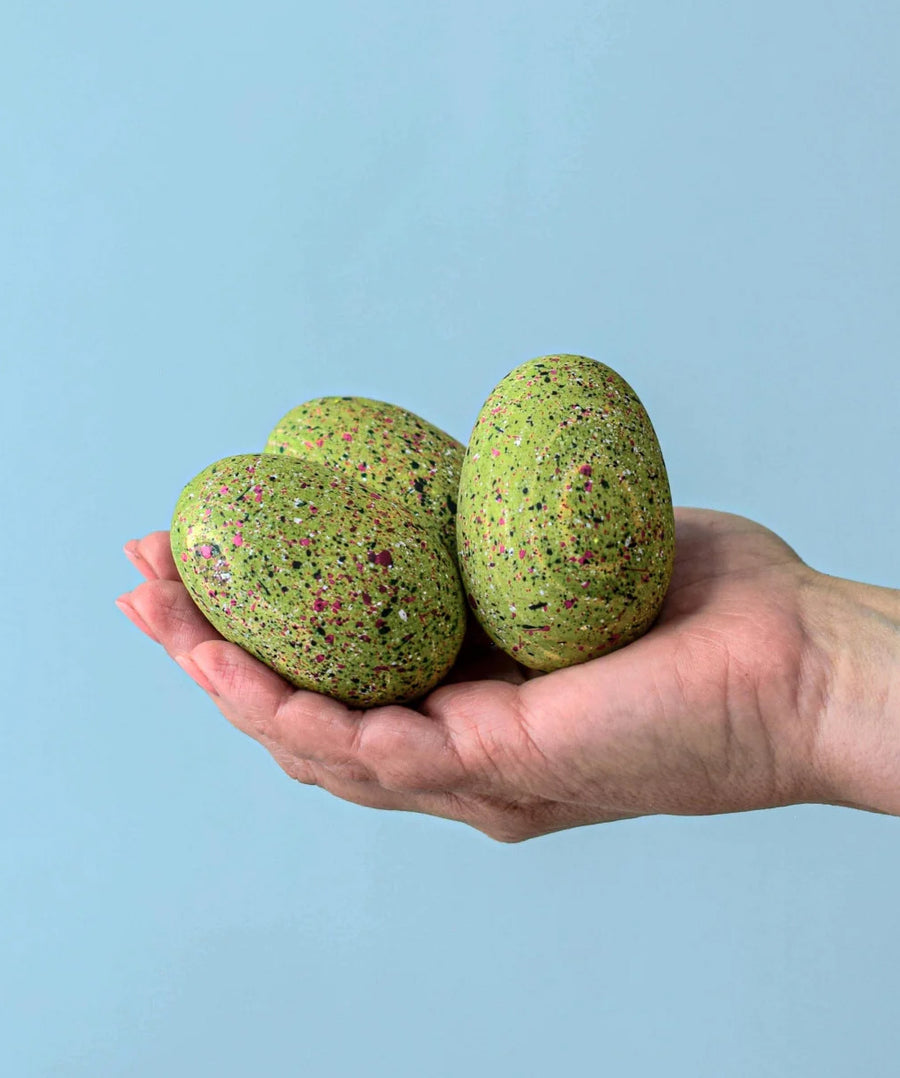 A close up of an adult holding the three bumbu t-rex eggs in their hand against a blue background. 