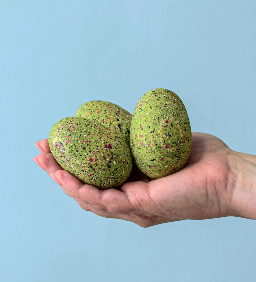 A close up of an adult holding the three bumbu t-rex eggs in their hand against a blue background. 