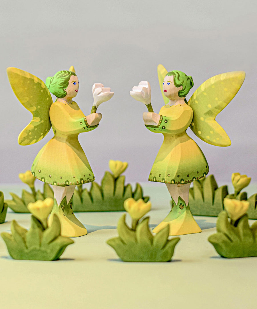 Two Bumbu woodland fairies curronded by yellow wooden flower figures. Each of teh fairies are holding small white flowers.