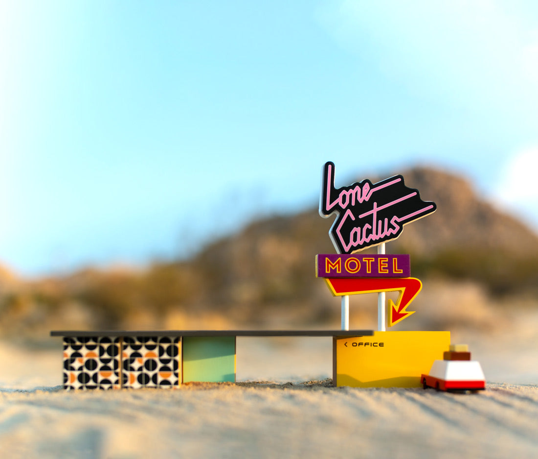 Close up of the Candylab wooden luggage wagon toy parked outside the wooden toy motel on a sandy surface