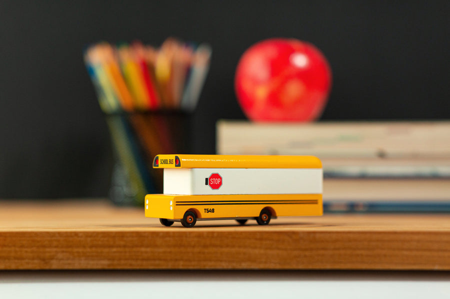 Candylab wooden bus vehicle toy on a wooden desk in front of some books and a red apple