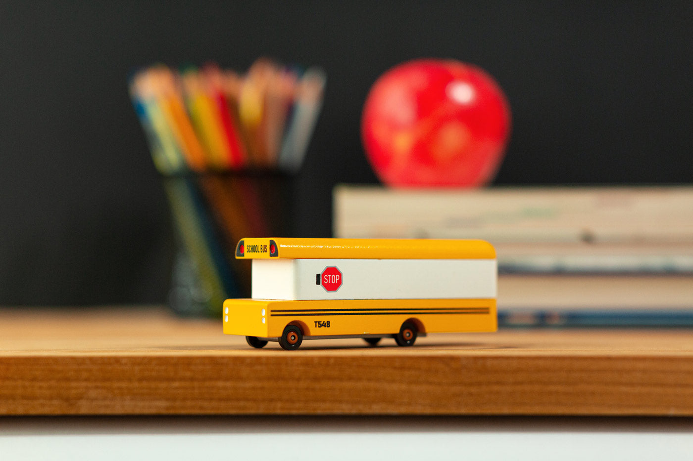 Candylab wooden bus vehicle toy on a wooden desk in front of some books and a red apple