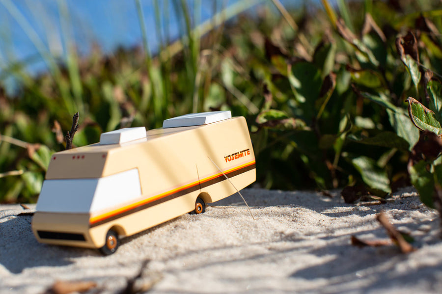 Close up of the Candylab wooden diecast campervan toy on some light sand in front of some green leaves