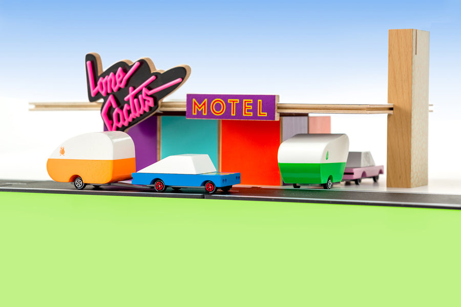 Candylab handmade wooden racing car and pink sedan toys towing wooden caravans into a toy motel model on a green and blue background