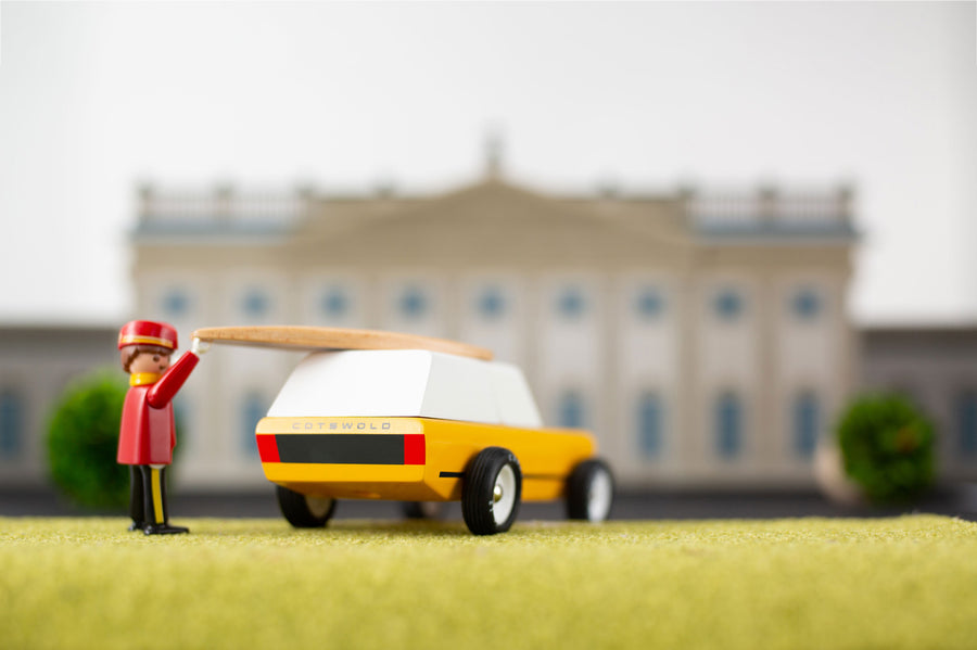 Toy figure putting a miniature surfboard on top of the candylab gold cotswold toy vehicle in front of a mansion model