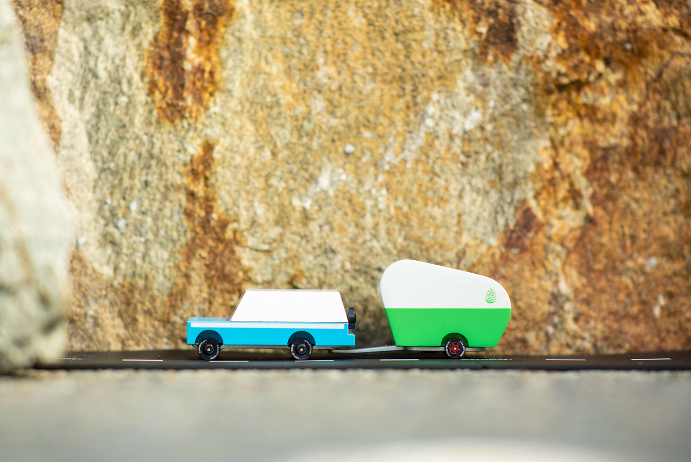 Candylab wooden mule car toy with a toy pinecone trailer attached on a rubber road in front of a large rock