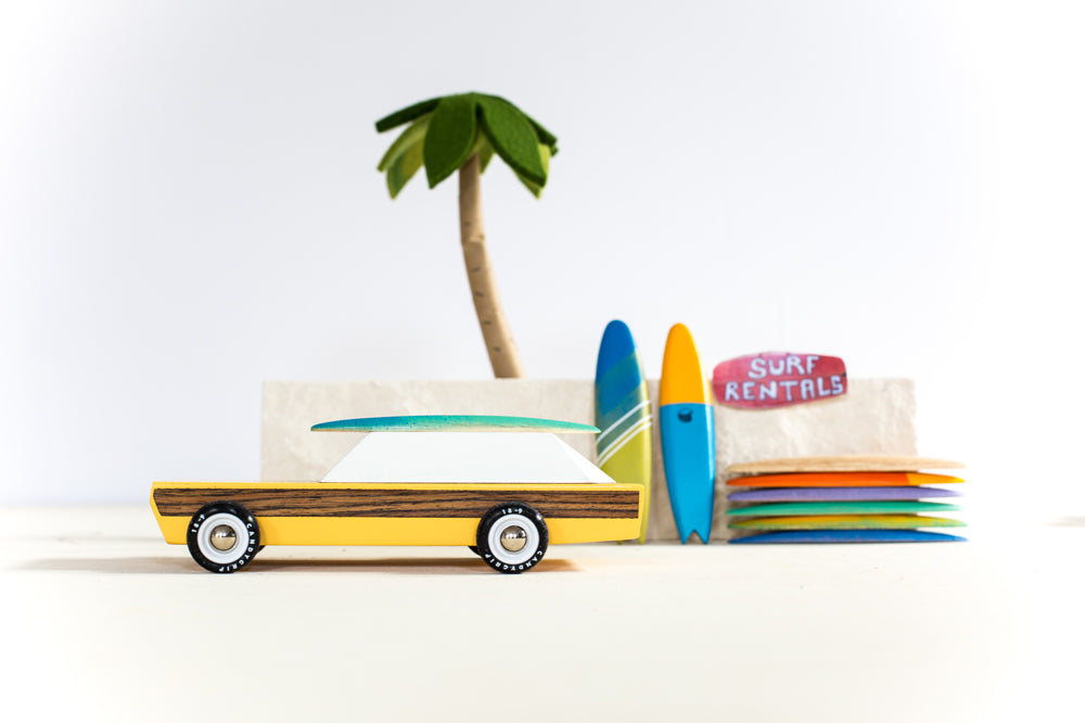 Candylab collectable yellow woodie car toy on a sandy background in front of some miniature surfboards and palm tree