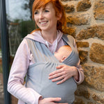 Woman leaning against a stone wall holding a baby in the close caboo lite baby carrier in the light grey alloy colour