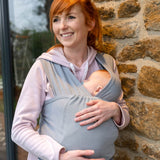 Woman leaning against a stone wall holding a baby in the close caboo lite baby carrier in the light grey alloy colour