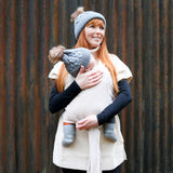 Woman stood in front of a metal wall holding a baby in the close caboo lite baby carrier in the blush cream colour