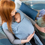 Woman laying on some cushions holding a baby in the close caboo lite baby carrier in the denim blue colour