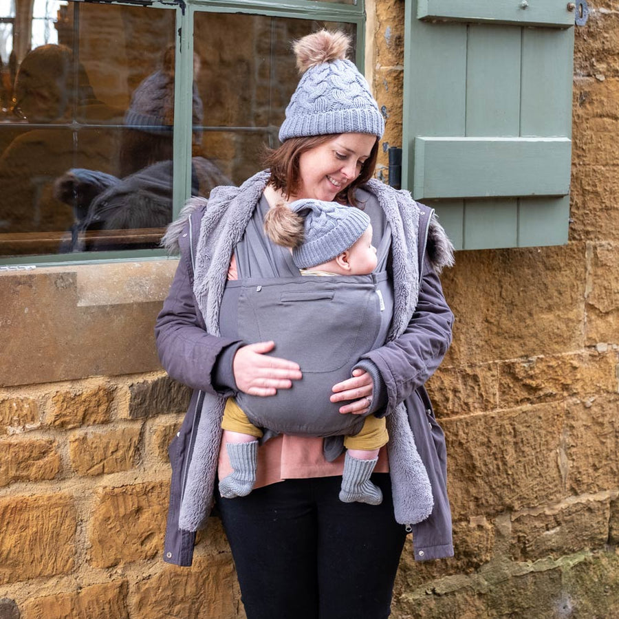 Woman stood in front of a stone wall holding her baby in the close caboo cotton blend baby carrier in the olive colour