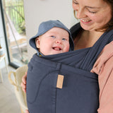 Woman stood in some woods wearing the Close caboo organic baby carrier in the blueberry blue colour