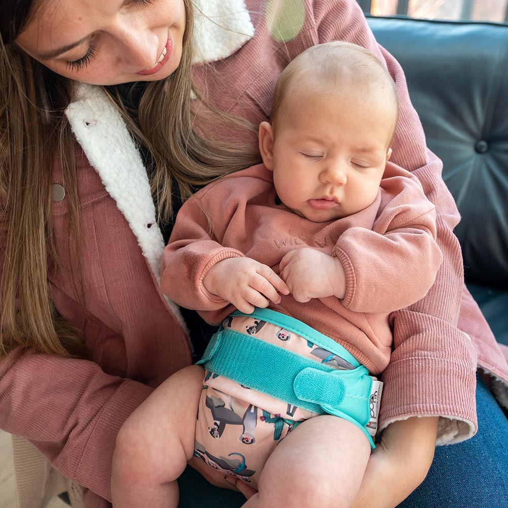 Sleeping baby wearing Pop-in light pink Ferret velcro Nappy all in one nappy with green trim details being held by a woman