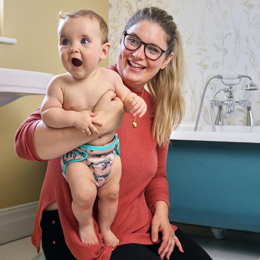 Surprised baby wearing Pop-in light pink Ferret popper Nappy all in one nappy with green trim details held by an adult