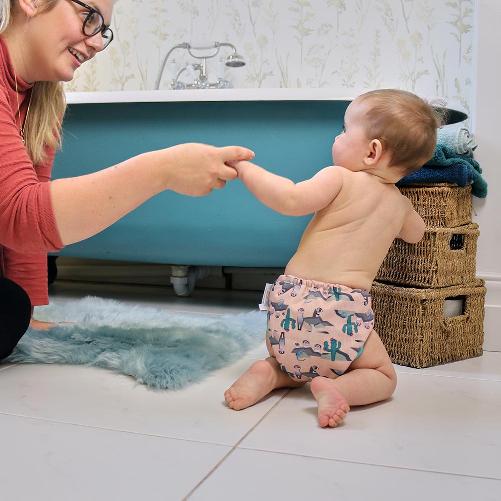 Baby wearing Pop-in light pink Ferret popper Nappy all in one nappy with green trim details with bath in background holding onto adults hand