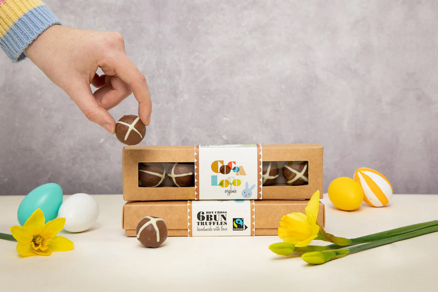 Cocoa Loco fairtrade hot cross bun easter chocolate truffles boxes and truffle being held in a persons fingers. On a white tables with colourful toys eggs