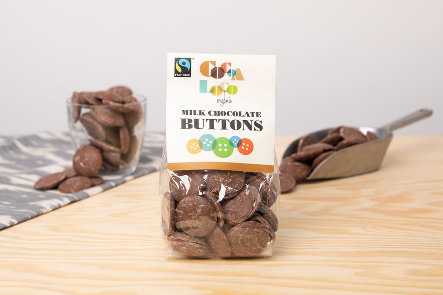 Bag of Cocoa Loco organic milk chocolate buttons on a wooden table in front of a glass cup and metal scoop filled with chocolate buttons