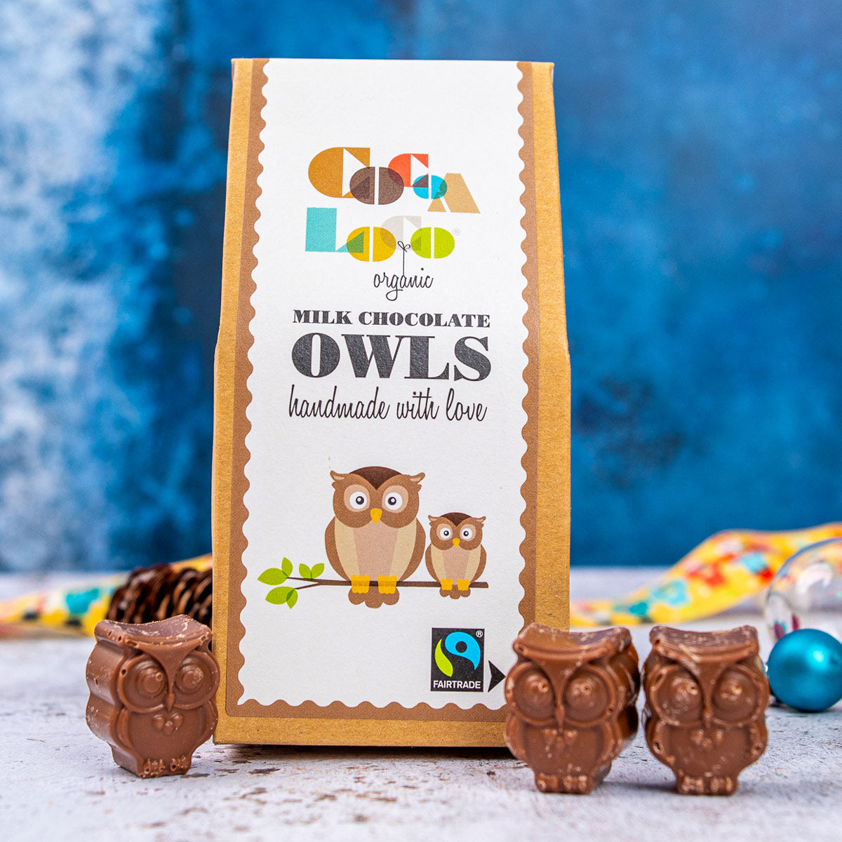 3 Cocoa Loco organic Fairtrade milk chocolate owls on a white background next to their cardboard packaging and some Christmas decorations