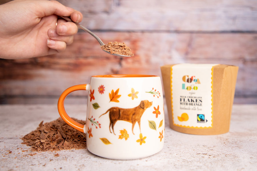 Close up of hand holding a spoon of Cocoa Loco Fairtade milk chocolate drinking flakes over a mug with a painted dog on the front