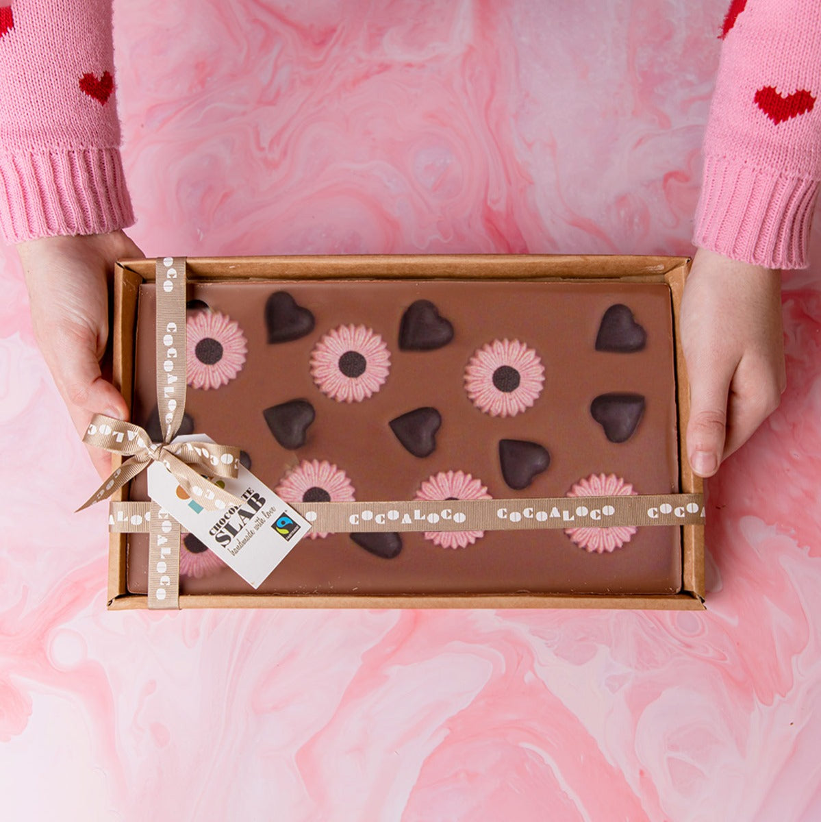 Close up of hands holding a Cocoa Loco organic milk chocolate hearts and flowers slab over a pink background