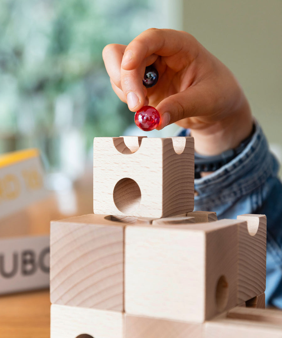 Close up of a child holding a red and black glass marble above a wooden Cuboro marble run set