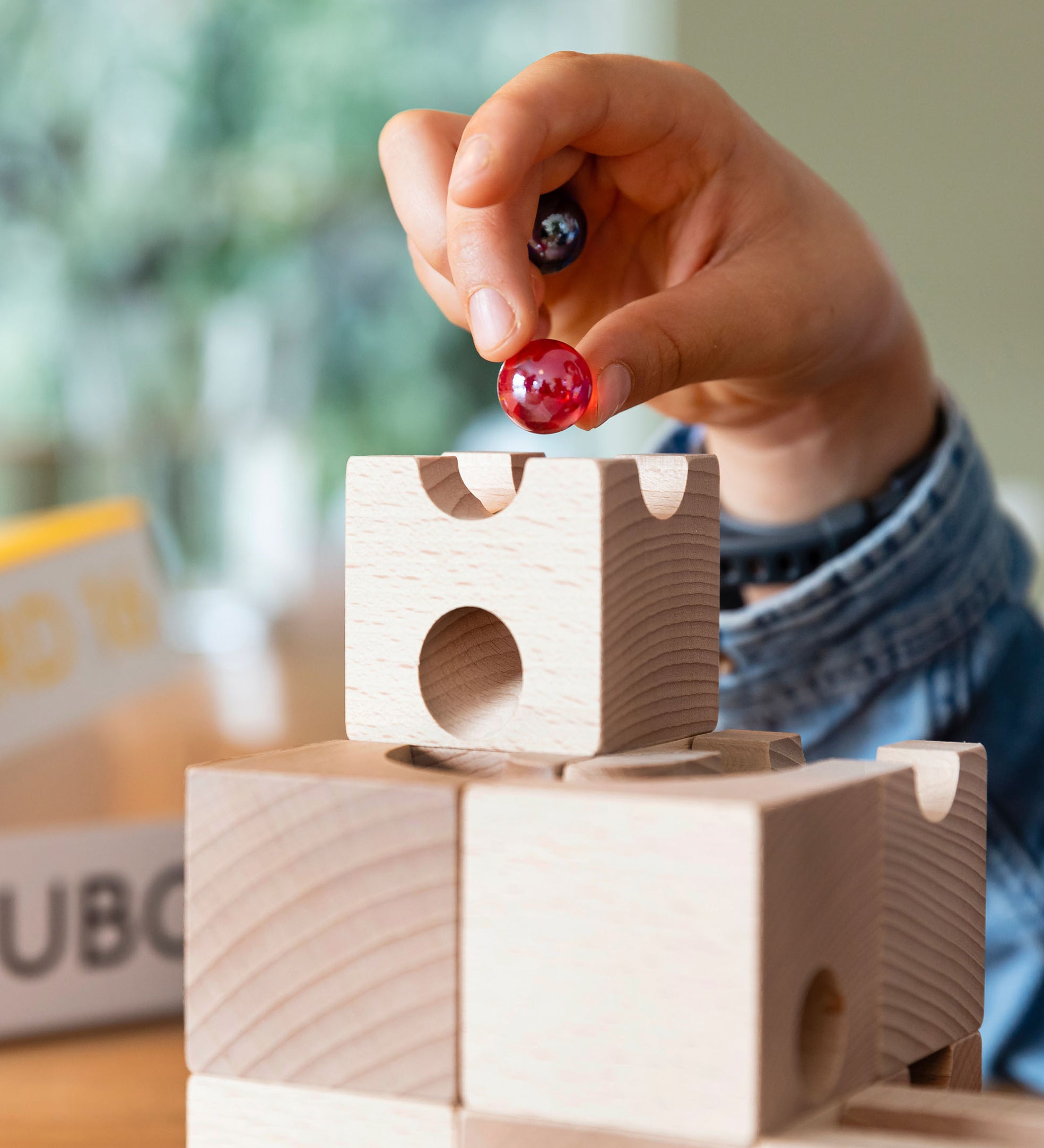 Close up of a child holding a red and black glass marble above a wooden Cuboro marble run set