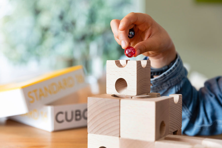 Close up of a child holding a red and black glass marble above a wooden Cuboro marble run set