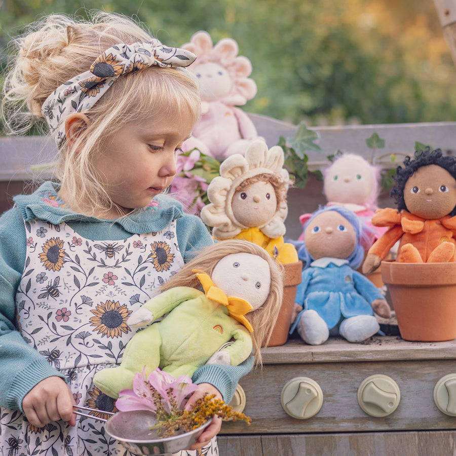 Olli Ella Dinky Dinkum Blossom Buds - Iris is sat on a garden bench surrounded by the rest of the blossom bud range with a child holding Sunflower doll in the foreground