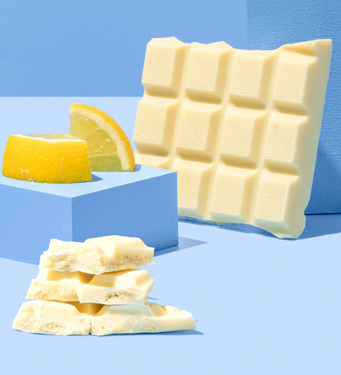 The Divine White Chocolate Lemon Cheesecake Dessert Bar shown unwrapped and broken up with a pile of the pieces shown at the front with pieces of lemon behind. 