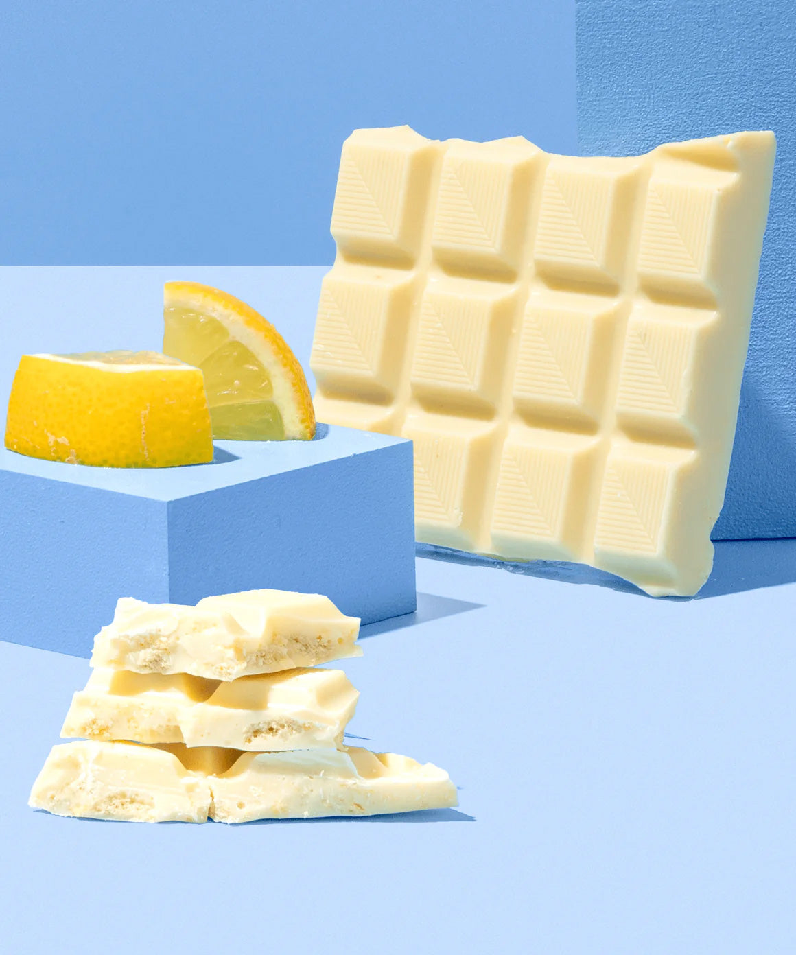 The Divine White Chocolate Lemon Cheesecake Dessert Bar shown unwrapped and broken up with a pile of the pieces shown at the front with pieces of lemon behind. 