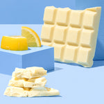 The Divine White Chocolate Lemon Cheesecake Dessert Bar shown unwrapped and broken up with a pile of the pieces shown at the front with pieces of lemon behind. 