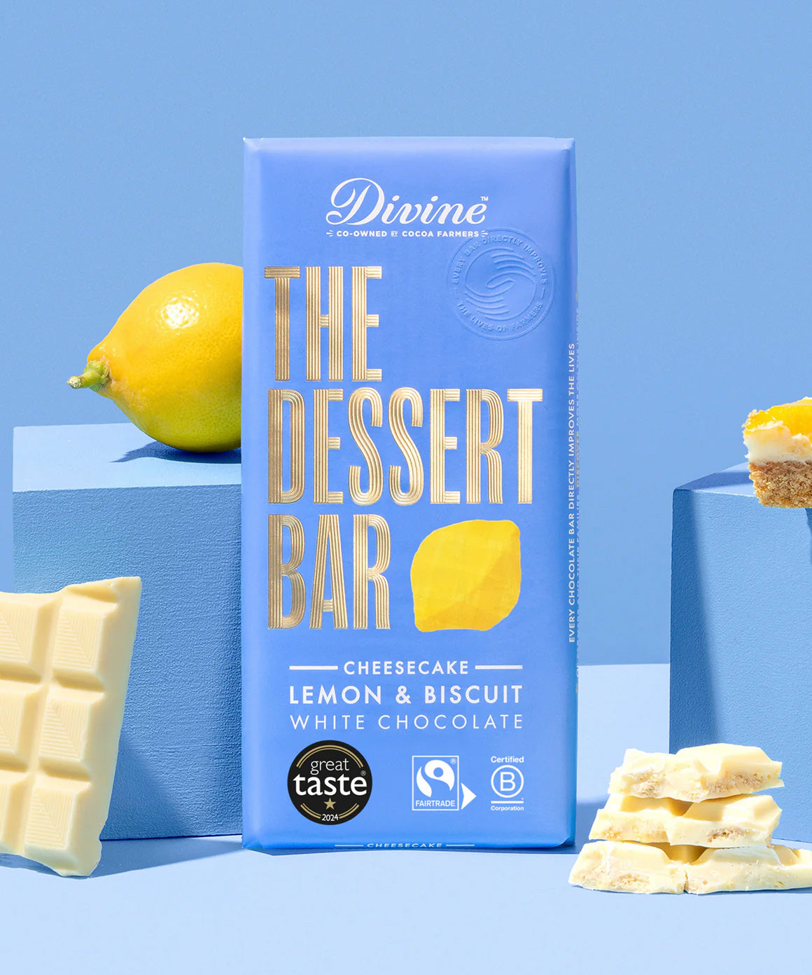 The The Divine White Chocolate Lemon Cheesecake Dessert Bar in it's wrapping. An unwrapped bar can be seen along with some lemons and a cheesecake on the sides. 