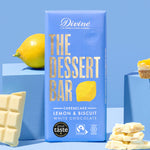 The The Divine White Chocolate Lemon Cheesecake Dessert Bar in it's wrapping. An unwrapped bar can be seen along with some lemons and a cheesecake on the sides. 