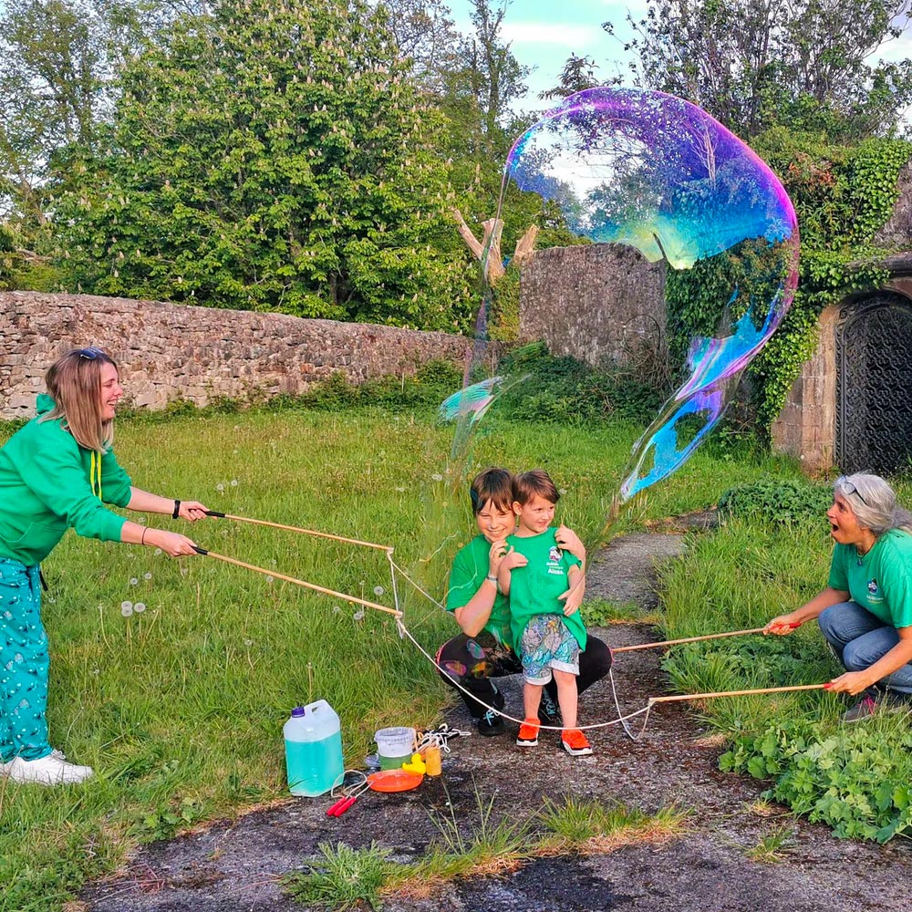 2 children crouched down on a path with two women blowing a Dr Zigs giant bubble around them