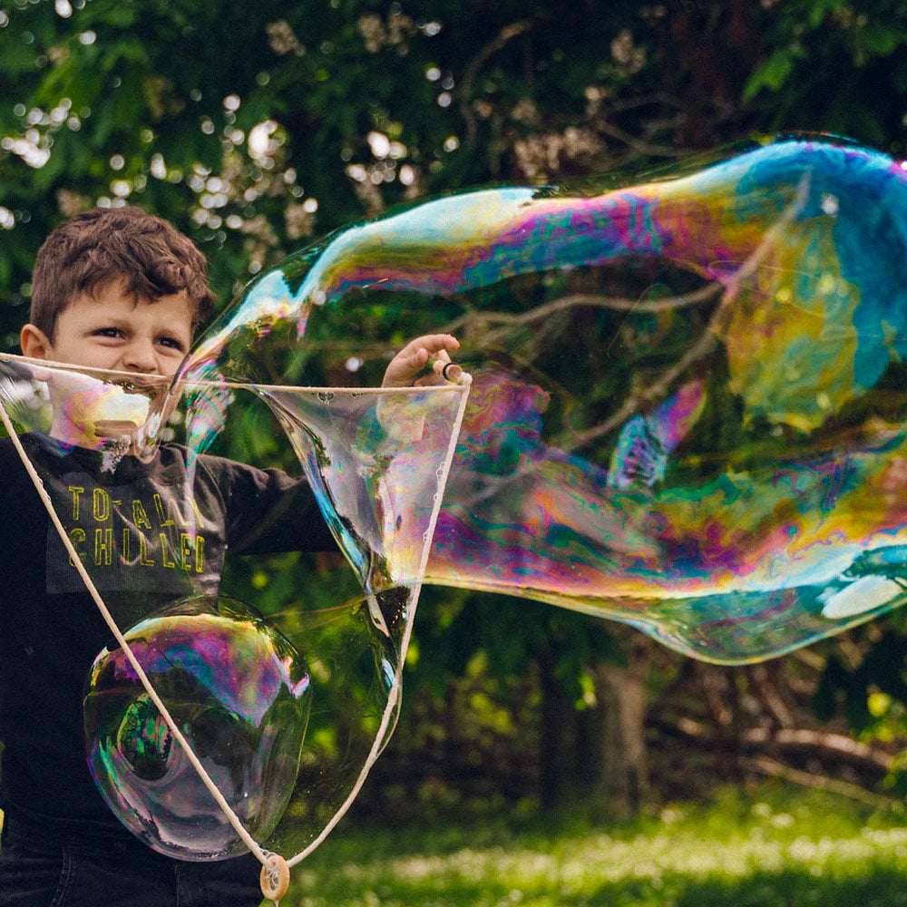 Close up of boy blowing a large bubble from the Dr Zigs wooden bubble travel kit wand in front of some trees