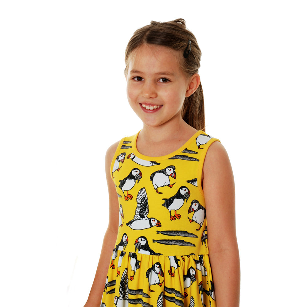 Girl stood on a white background wearing the DUNS Sweden organic cotton sleeveless gather skirt dress in the yellow puffin print