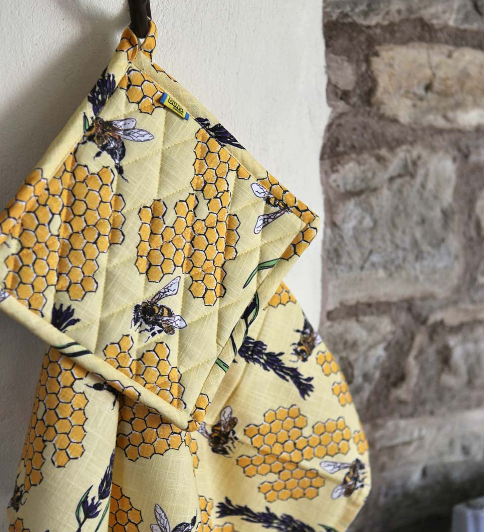 DUNS Sweden organic cotton linen pot holder and tea towel in the bee yellow print hanging from a metal hook in front of a stone wall